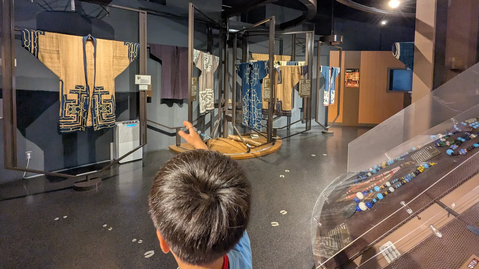 Child points toward a display of Ainu clothing and textiles at the Sapporo Ainu Culture Promotion Center in Hokkaido, learning about Indigenous culture.