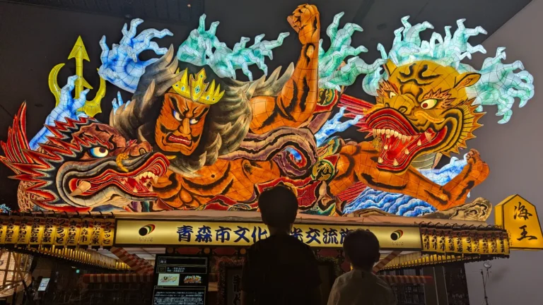 Two kids gaze up at a giant illuminated Nebuta Festival float inside Nebuta Museum Wa Rasse in Aomori, Japan, a top family attraction filled with color and culture.