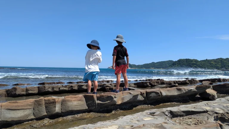 Luca & Nico standing on the Devil’s Washboard rock formations at Aoshima Beach, looking out at the waves on a sunny day.