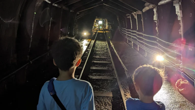 Two boys stand near railway tracks leading out of the Ashio Copper Mine Museum tunnel, light shining at the end of the mine passage.