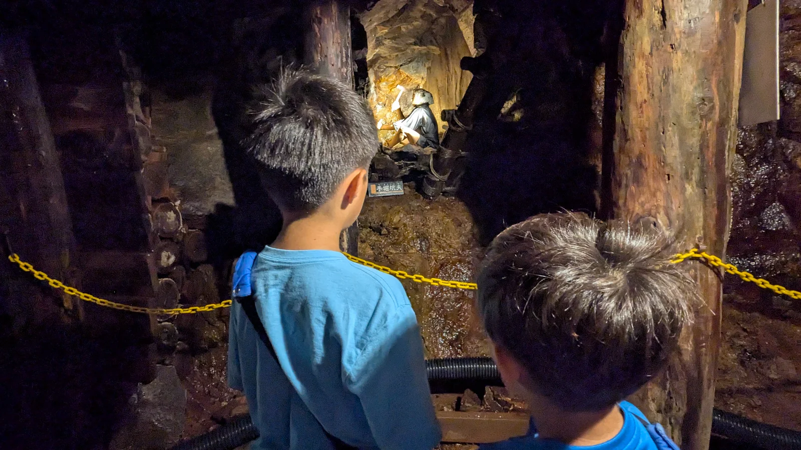 Children stand in a dimly lit tunnel at the Ashio Copper Mine Museum, watching a miner mannequin chiseling rock by lantern light.