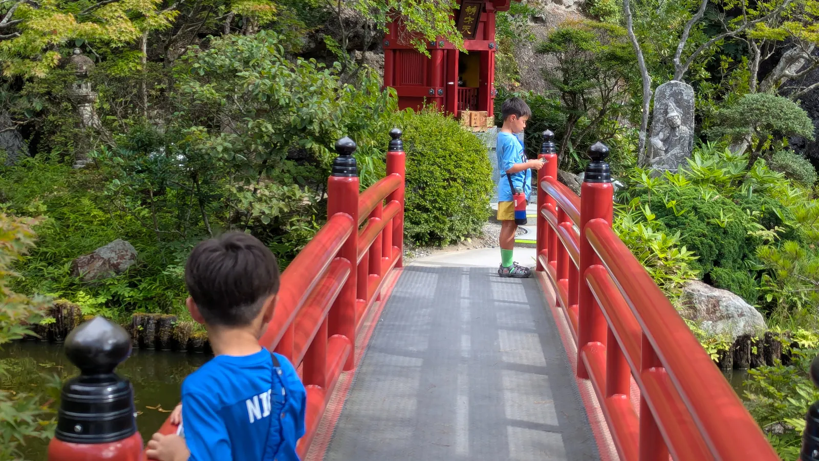 Two children cross the bright red bridge leading to Bentendo Hall at Oya Temple, surrounded by lush green trees and stone carvings.