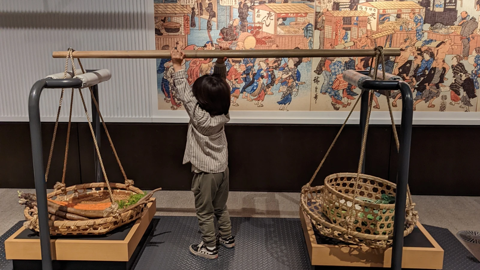 Child trying a hands-on Edo-Tokyo Museum exhibit, lifting traditional baskets used for carrying goods in old Tokyo.