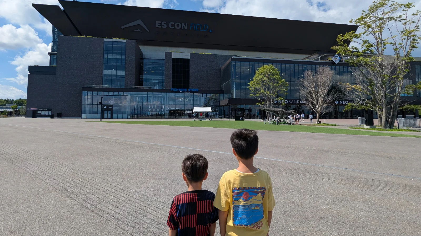 Two kids stand outside Es Con Field Hokkaido near the Fighters Store entrance, looking up at the modern stadium design on a sunny day.