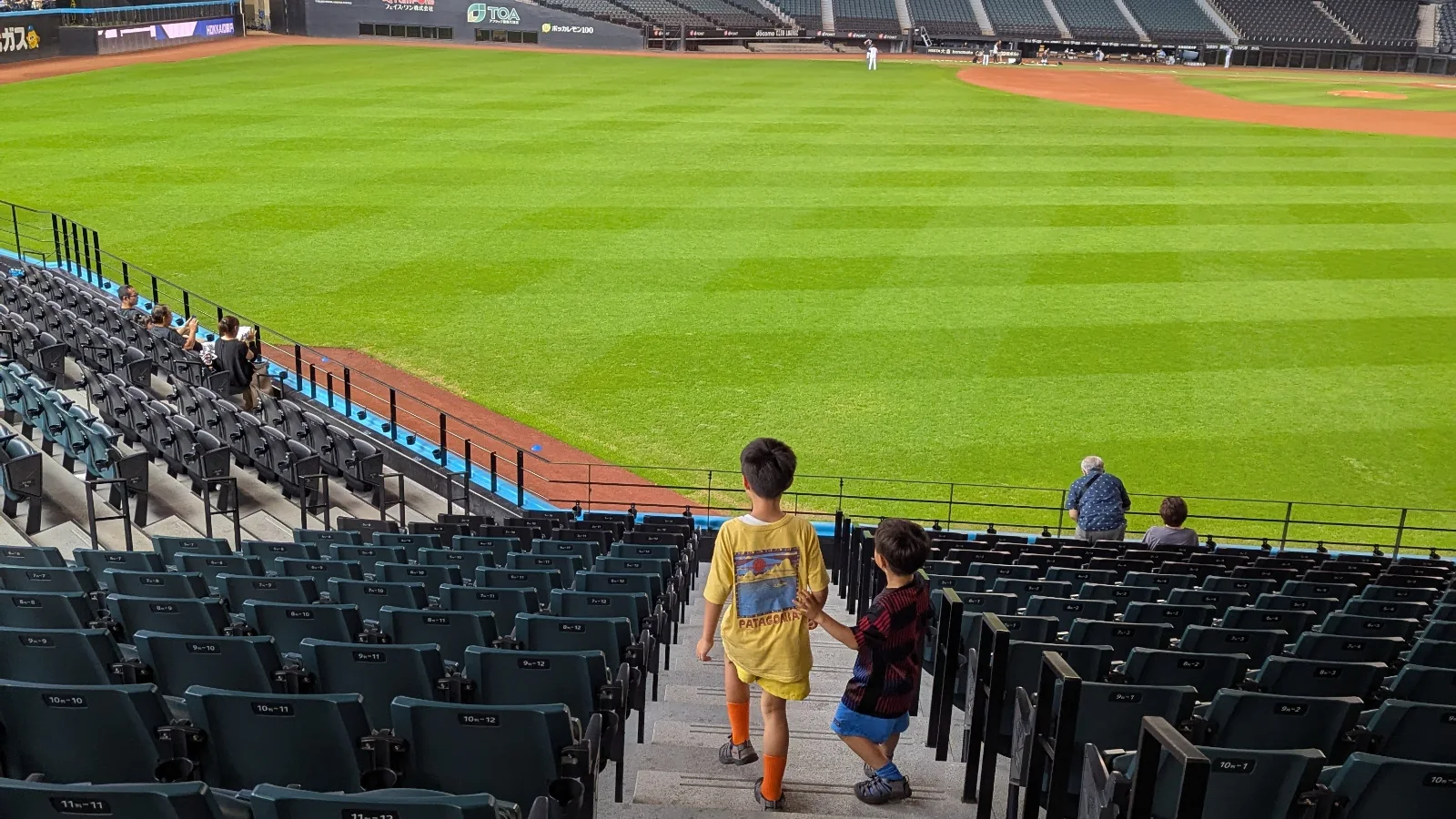 Two children walk down the stands toward the outfield at Es Con Field Hokkaido, exploring the open stadium on a family visit.