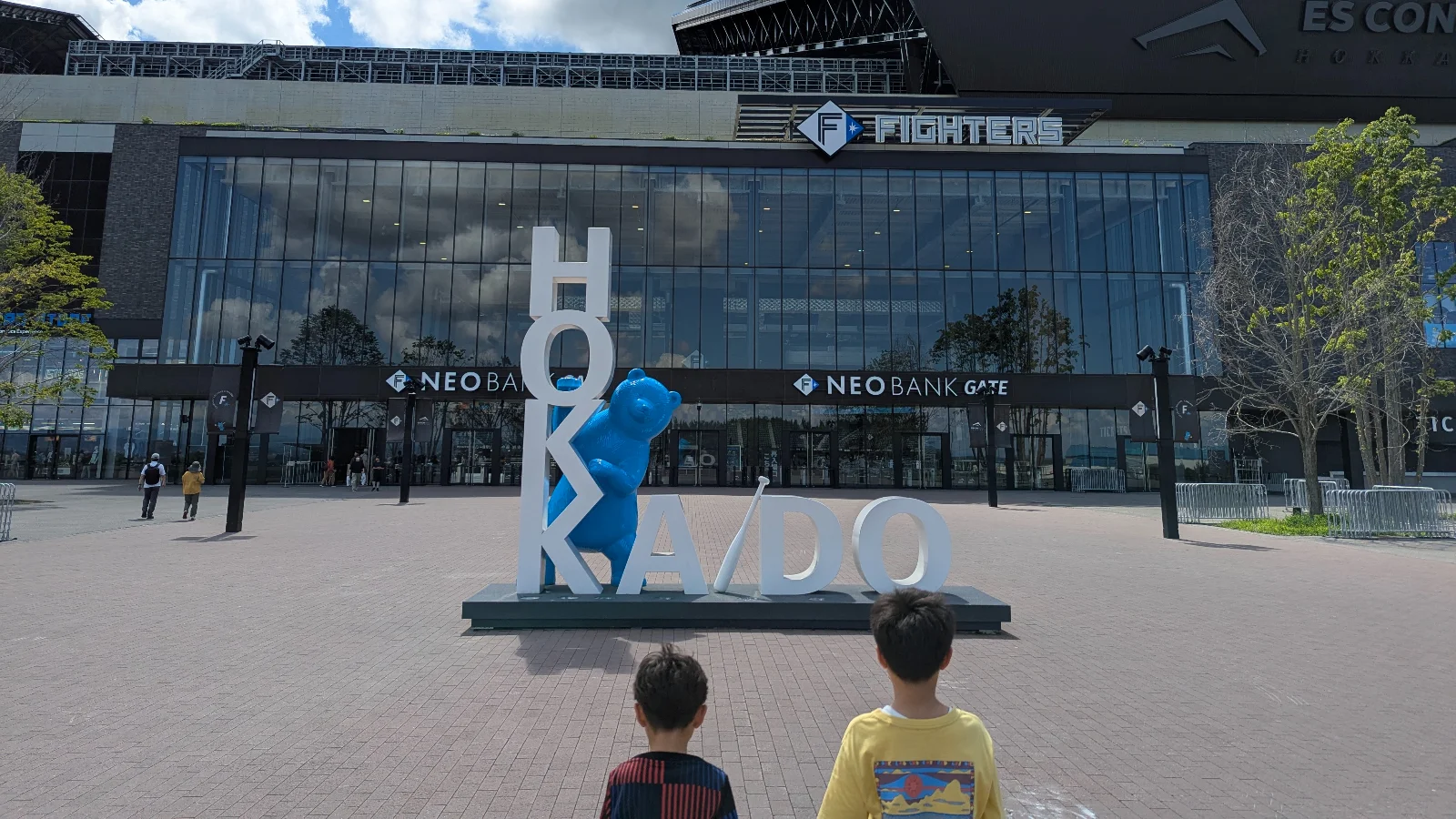 Two kids pose by the blue bear and HOKKAIDO sign in front of Es Con Field Hokkaido, a popular photo spot at F Village.