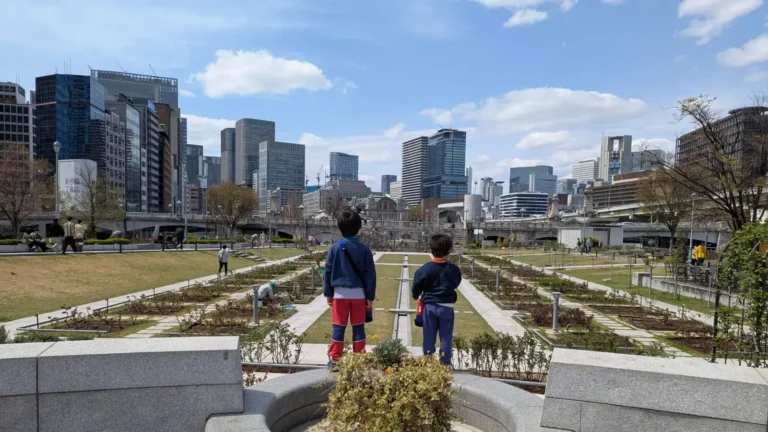 Luca and Nico explore a beautiful rose garden in the heart of Osaka, Japan, with a backdrop of towering skyscrapers. Discover family-friendly adventures in modern cities through the eyes of curious young travelers.