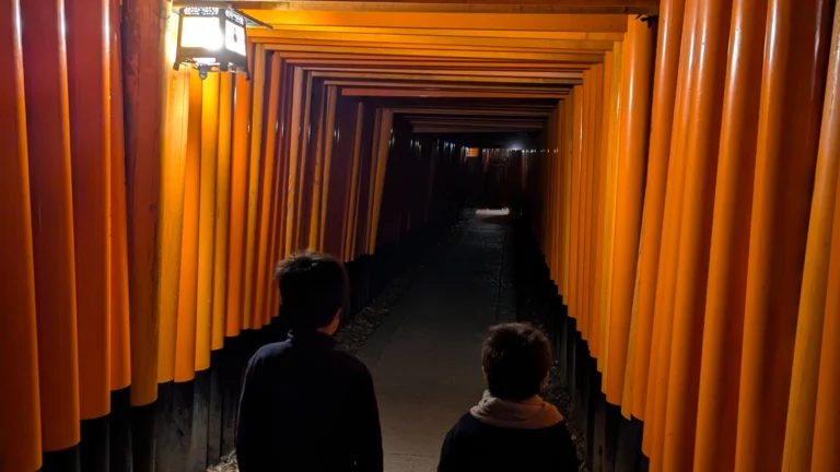 Two children walking through the endless red torii gates of Fushimi Inari Taisha in Kyoto, Japan, during a family visit. The vibrant pathway of Shinto gates creates a tunnel of color that captures the spirit of this iconic shrine, perfect for families exploring Kyoto with kids.