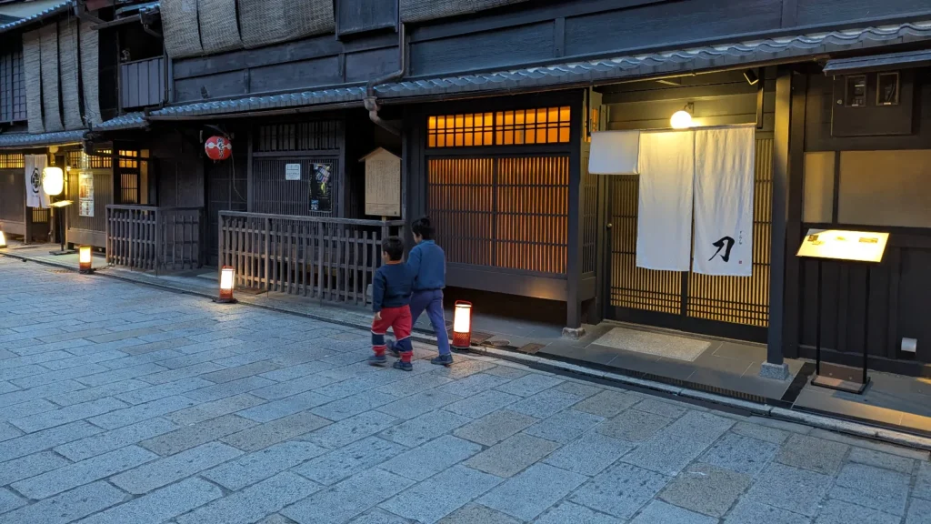 Luca & Nico walking along a lantern-lit street in the historic Gion district of Kyoto, surrounded by traditional wooden teahouses.