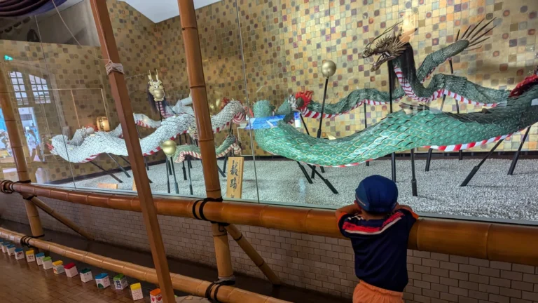 Young child in blue cap and orange shorts looks at a colorful dragon festival float display inside Glover Garden in Nagasaki, Japan.