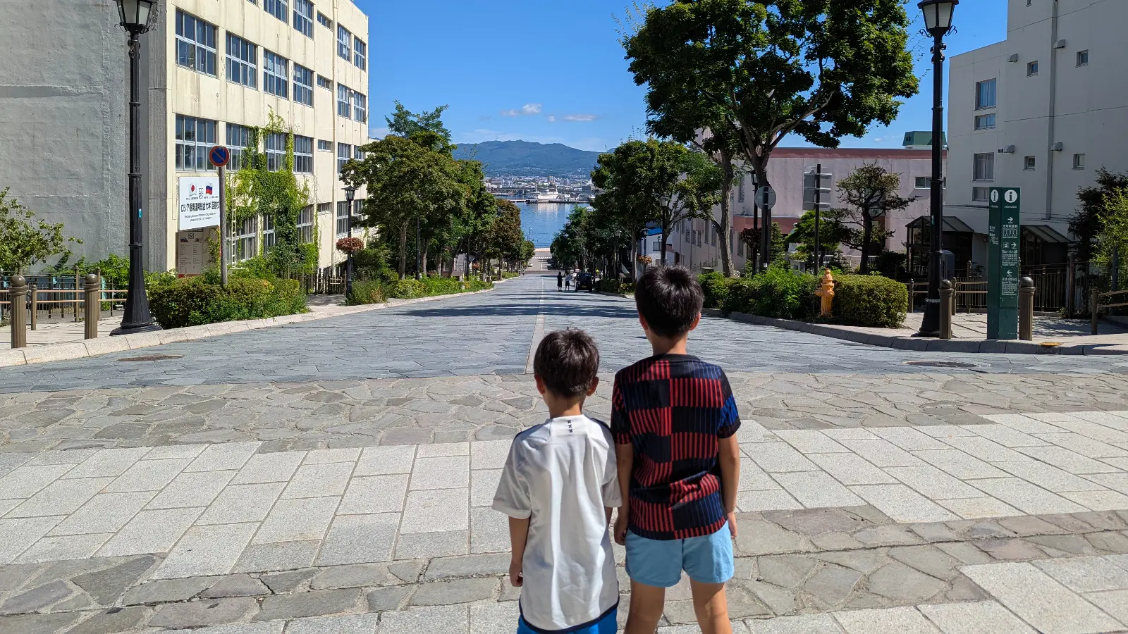 Two children look down the famous Hachiman-Zaka Slope toward Hakodate Harbor under blue skies, framed by trees and classic street lamps.