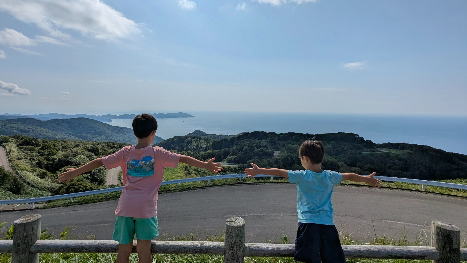 Two children stretch their arms wide at a scenic lookout over the Hakkōda Mountains and sea in Aomori, Japan, enjoying a family outdoor adventure.