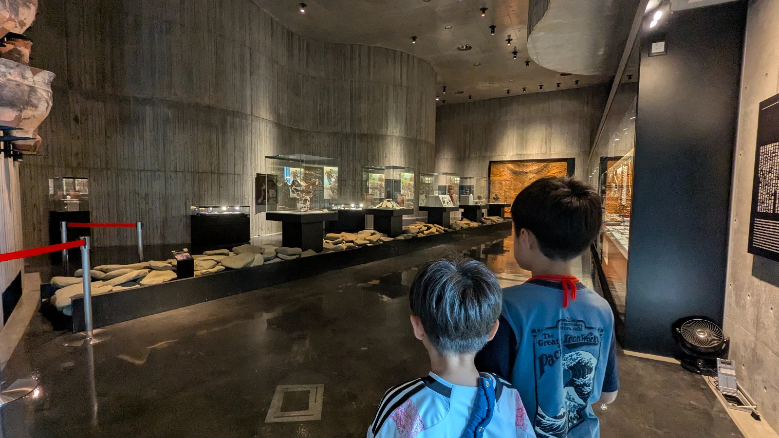 Two children explore the dimly lit exhibit hall at Hakodate Jomon Culture Center, filled with ancient artifacts and display cases.