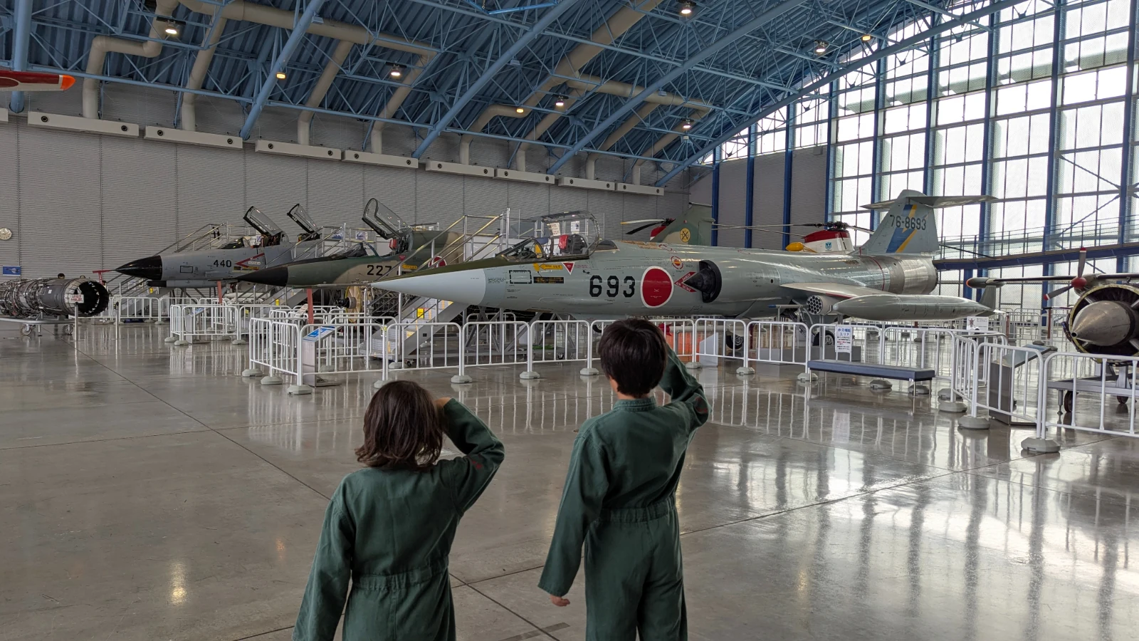 Luca and Nico, in green flight suits, salute a row of jet fighters at the JASDF Air Park in Hamamatsu.