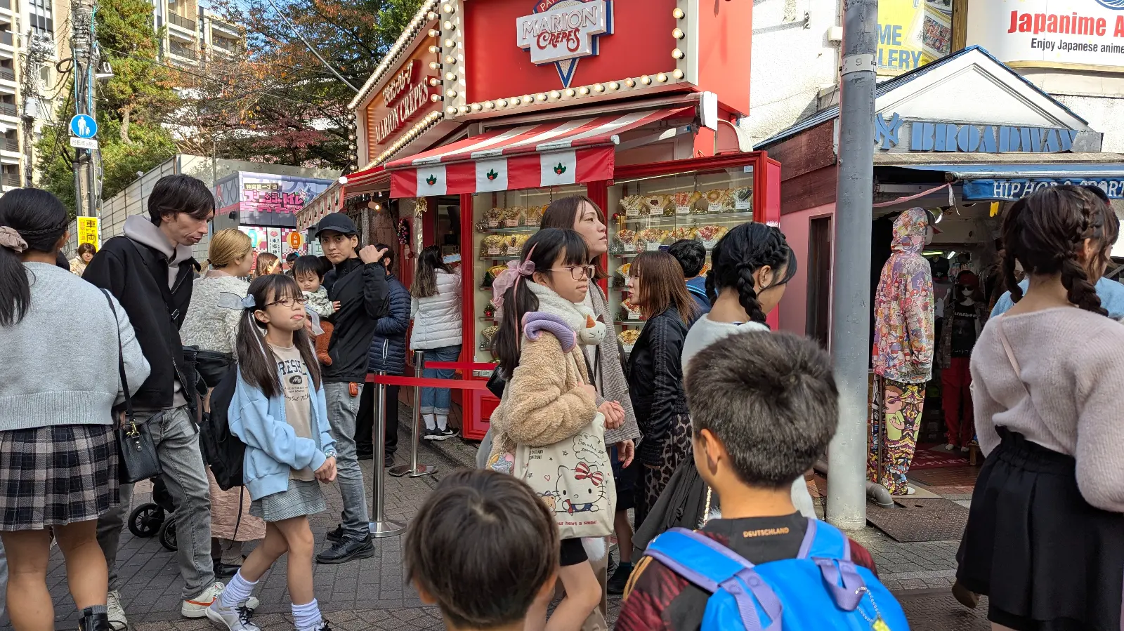 Crowds lining up at the famous Marion Crêpes shop on Takeshita Street in Harajuku, with two children walking through the busy food-lined alley.