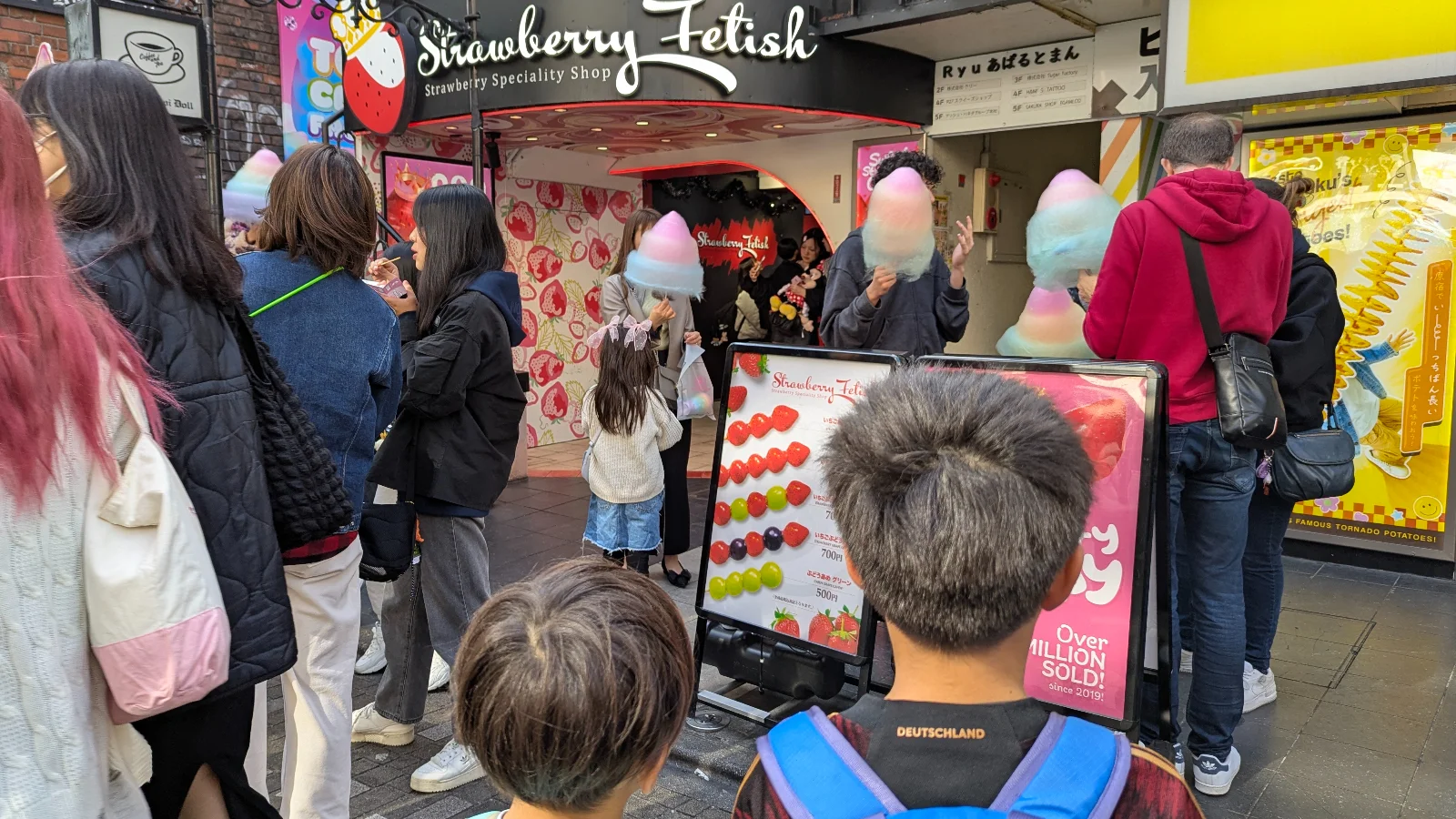 Visitors enjoying rainbow cotton candy in front of the Strawberry Fetish shop on Takeshita Street in Harajuku, with two children watching the colorful scene.