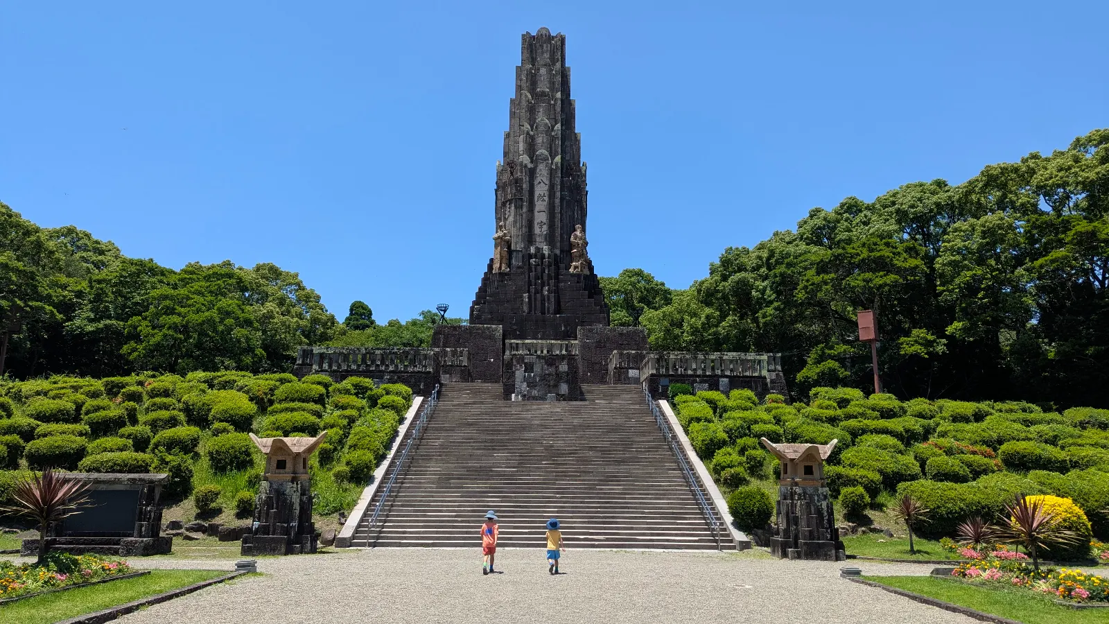 Luca & Nico walking toward the Peace Tower at Heiwadai Park, surrounded by neatly trimmed bushes and bright blue skies.