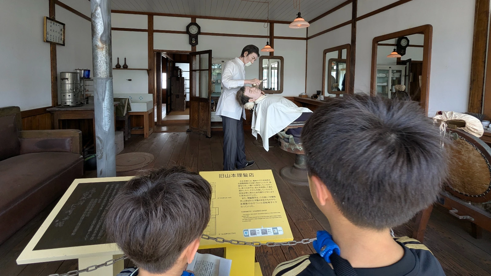 Children look into an old barbershop exhibit at the Historical Village of Hokkaido where mannequins show a traditional haircut scene.