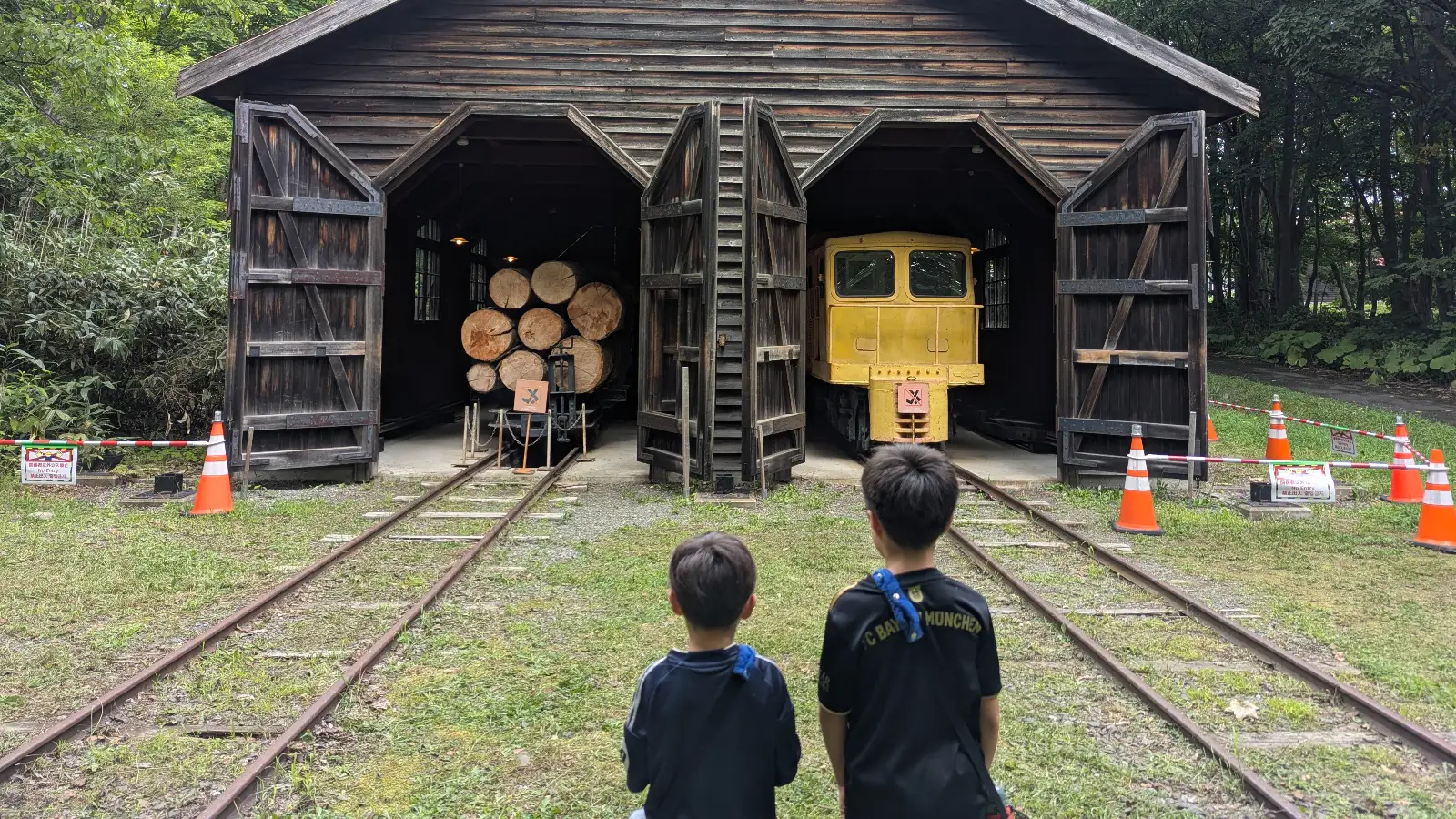 Two children stand in front of a wooden train shed at the Historical Village of Hokkaido in Sapporo, gazing at a vintage yellow locomotive loaded with timber logs.