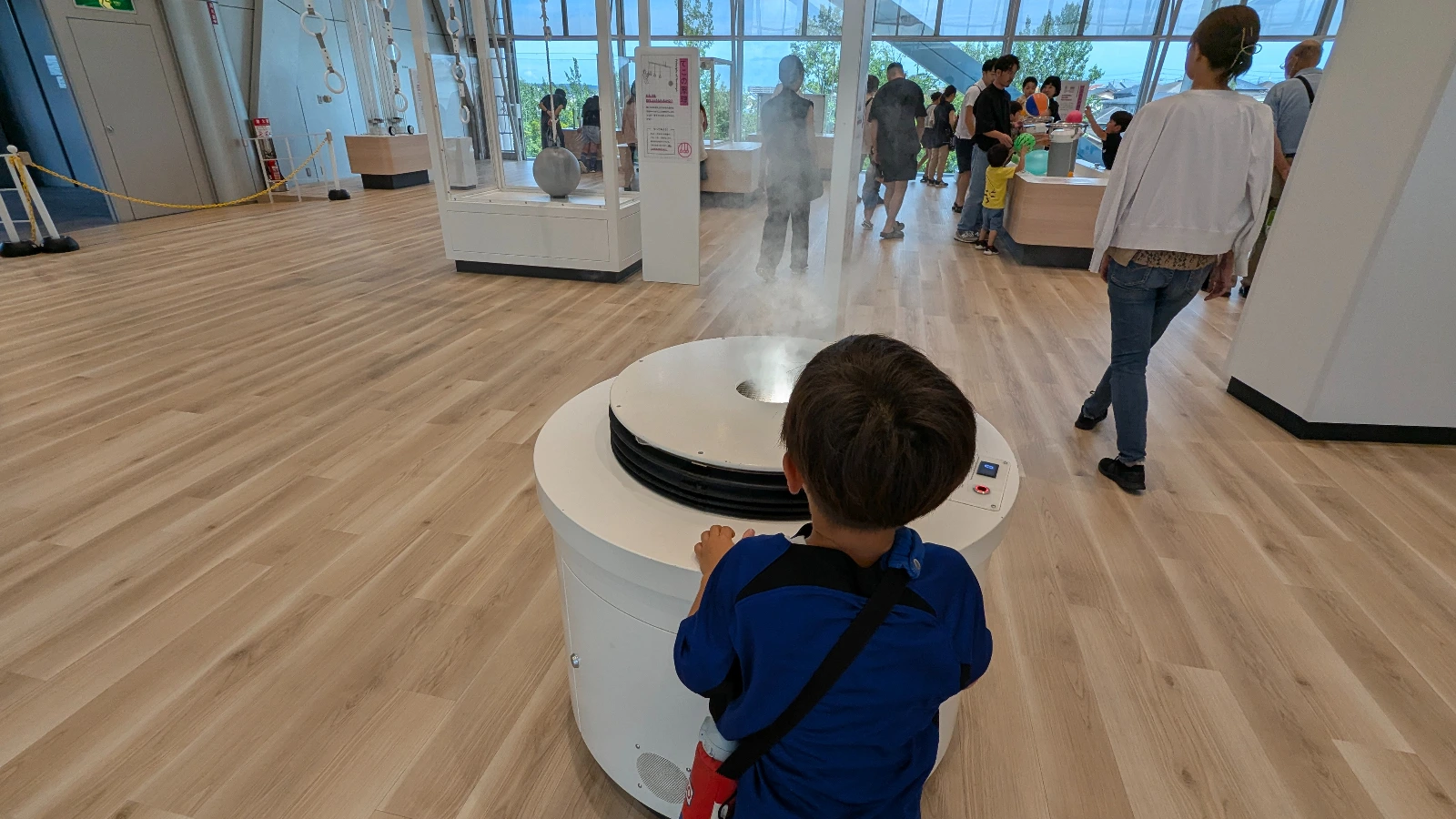 A young boy experiments with air flow at the Hokushu Sendai City Science Museum, watching mist rise through a circular air display inside the science hall.