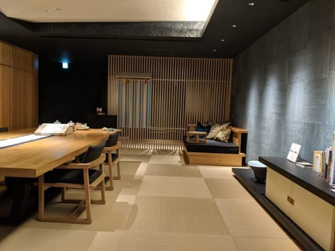 A serene and stylish Japanese tea lounge featuring tatami flooring, a wooden table with chairs, a cushioned seating area, and warm ambient lighting at Hoshinoya Tokyo.