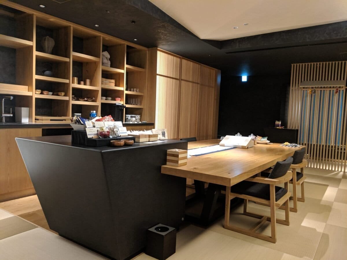 A cozy tea room with modern Japanese wooden shelving, a display of tea, sake, and snacks, and a spacious table for gatherings in a tatami room setting.