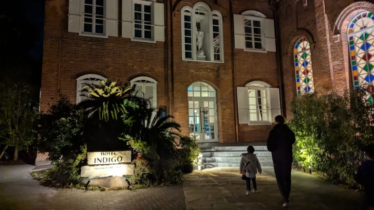 A family walks toward the lit entrance of Hotel Indigo Nagasaki at night, framed by red brick walls and colorful stained-glass windows.