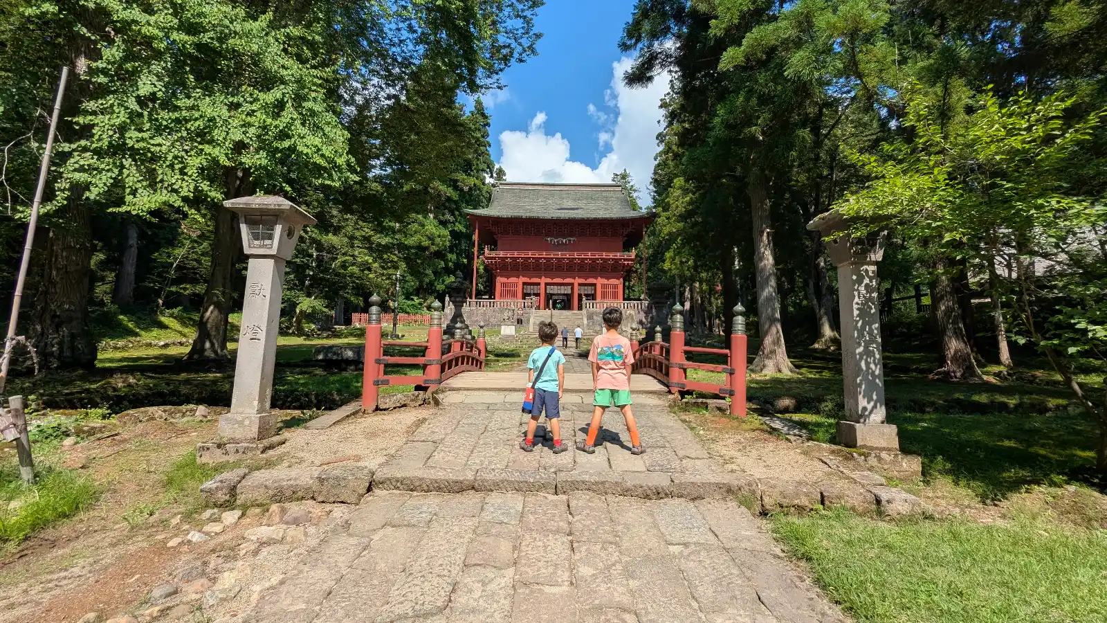 Two kids walk toward the main gate of Iwakiyama Shrine surrounded by towering cedar trees in Aomori, Japan, a peaceful and family-friendly cultural site.