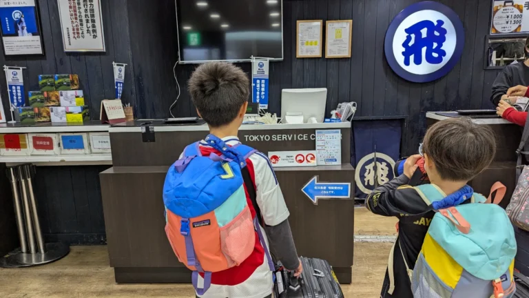 Two young kids with backpacks and suitcases standing at a Japan luggage delivery counter, using takuhaibin service before traveling, Tokyo station.