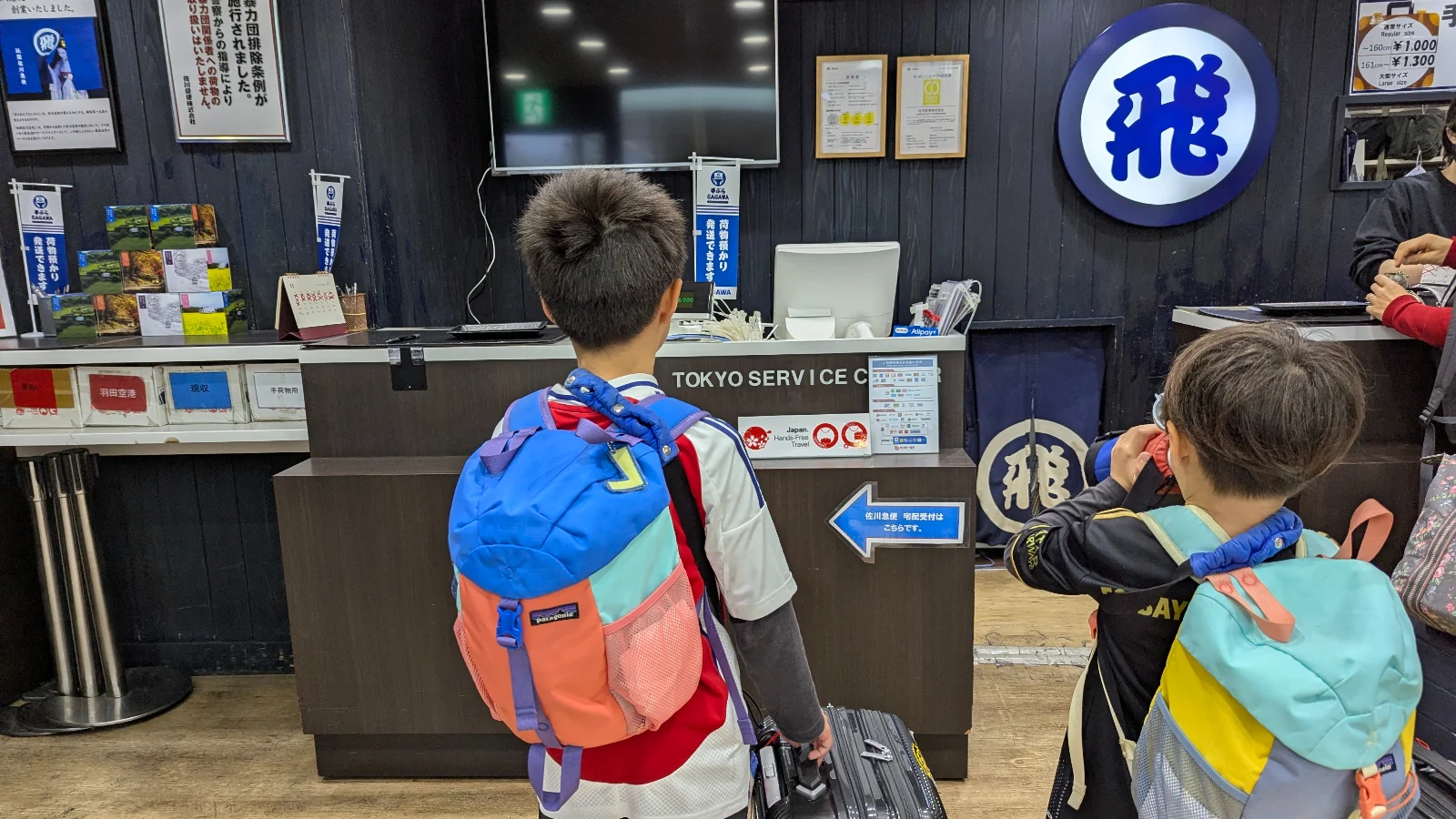 Two young kids with backpacks and suitcases standing at a Japan luggage delivery counter, using takuhaibin service before traveling, Tokyo station.