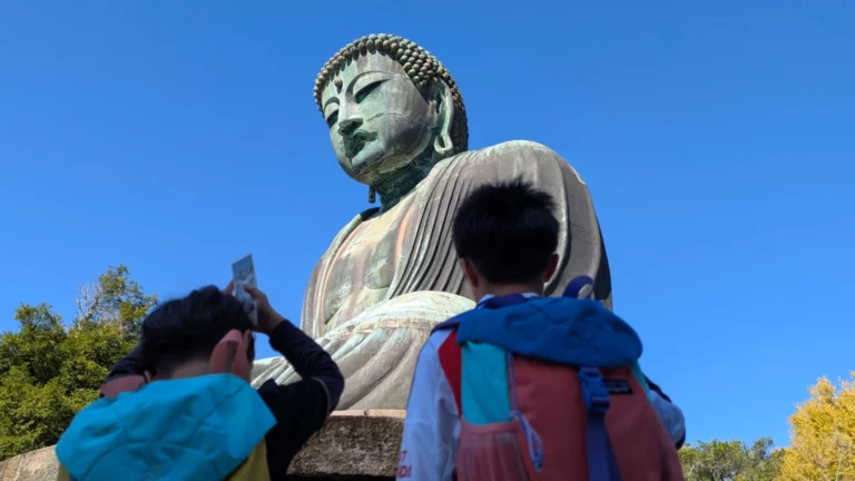 Two children with backpacks looking up at the Kamakura Great Buddha statue under a bright blue sky.