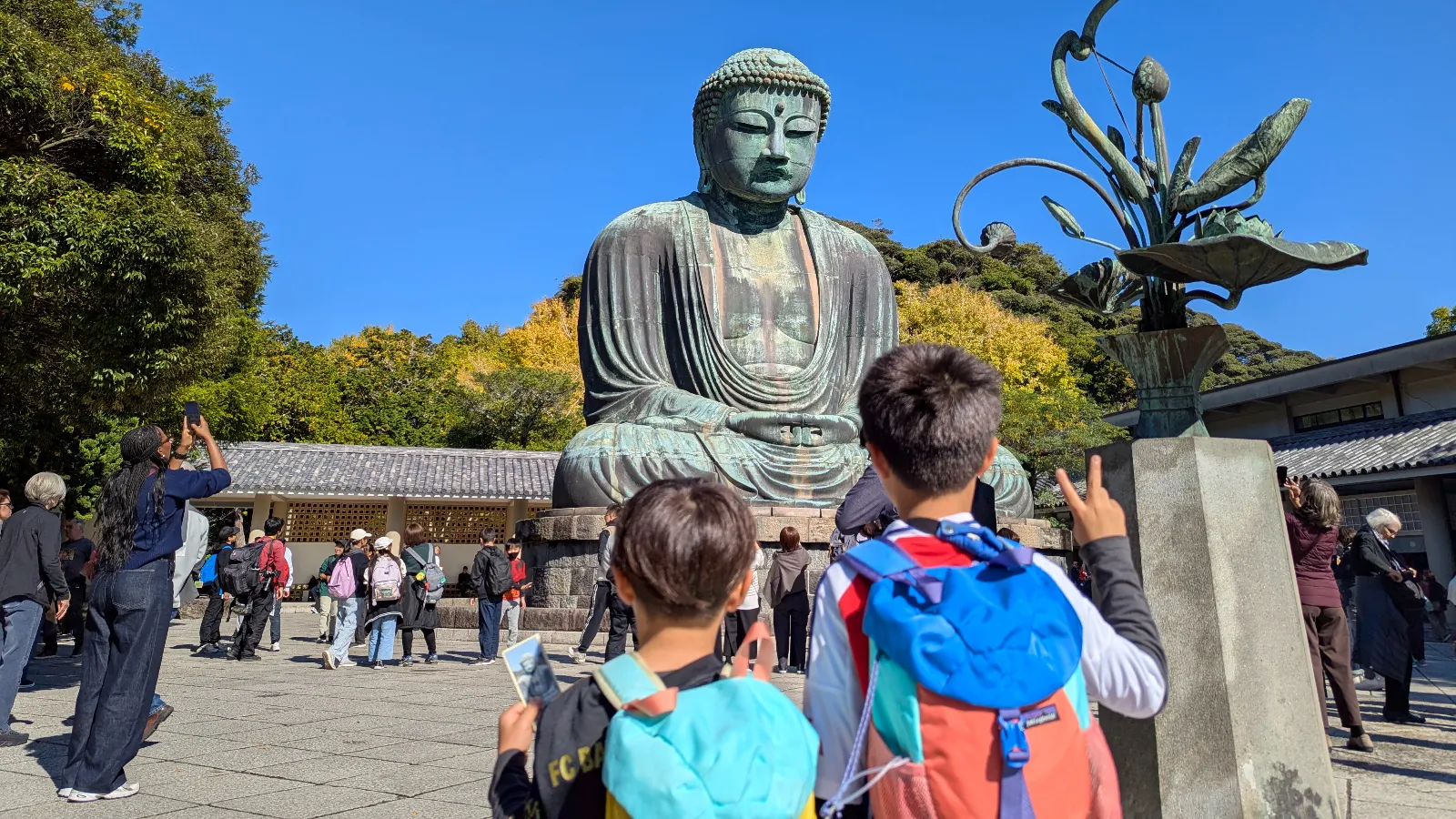 Two children standing before the Great Buddha of Kamakura at Kotoku-in Temple on a bright, sunny day.