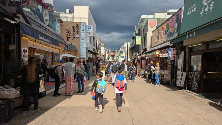 Children walking down Komachi Street in Kamakura, surrounded by shops, snacks, and crowds in the lively shopping district.