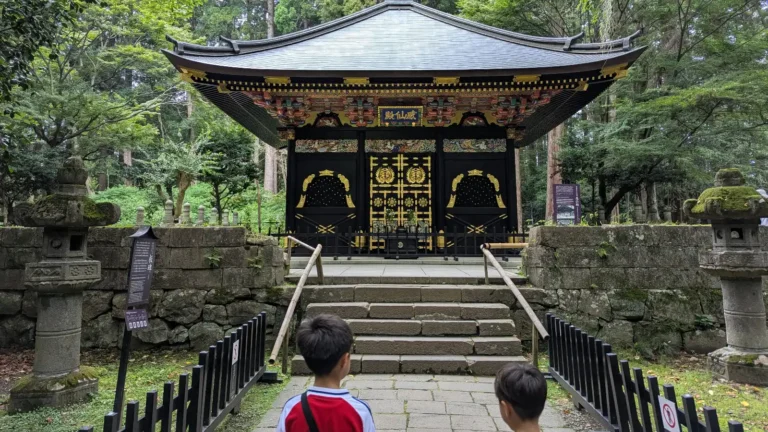 Two children look up at Kansenden, one of the ornate secondary mausoleums within the Zuihoden complex in Sendai, Japan, surrounded by cedar forest and stone lanterns.