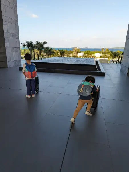 Luca & Nico pull rolling suitcases at a luxury resort entrance in Miyakojima, with a view of the Irabu Ohashi Bridge and ocean in the background, symbolizing family arrival and adventure.
