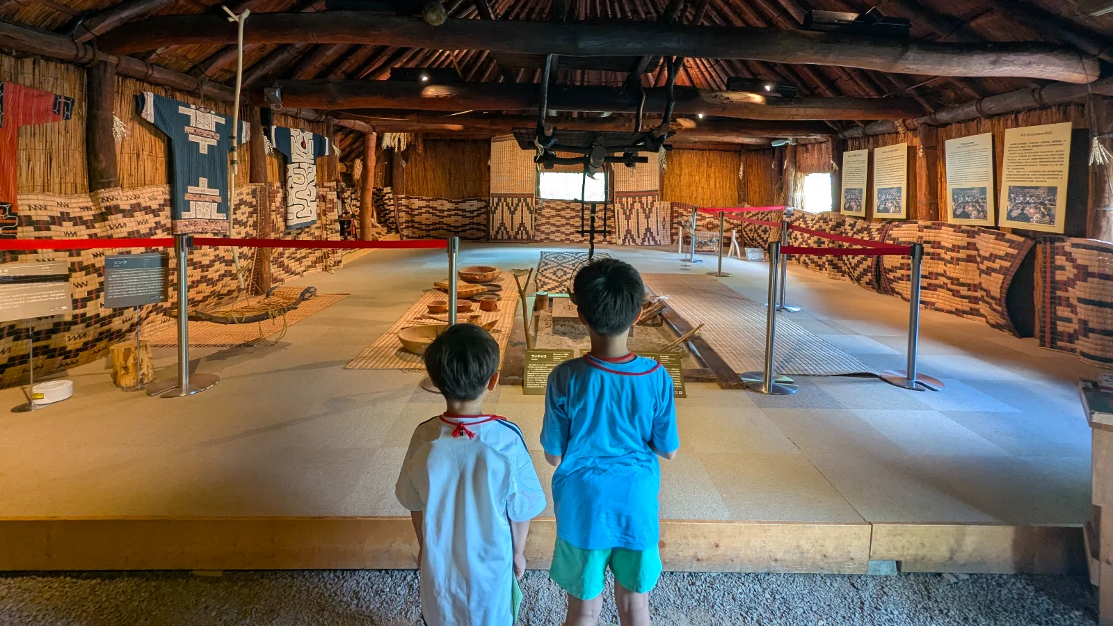Two children explore the interior of a reconstructed Ainu house at the Sapporo Ainu Culture Promotion Center, observing traditional tools and crafts.