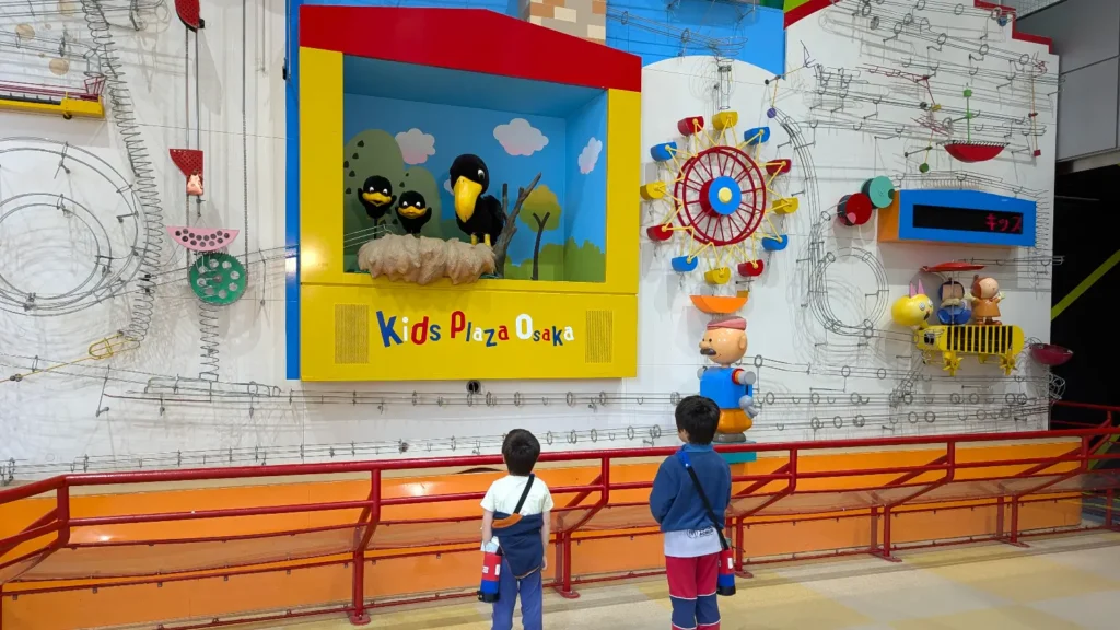 Luca and Nico watching a mechanical wall display at Kids Plaza Osaka, filled with colorful contraptions and moving parts that bring science to life.