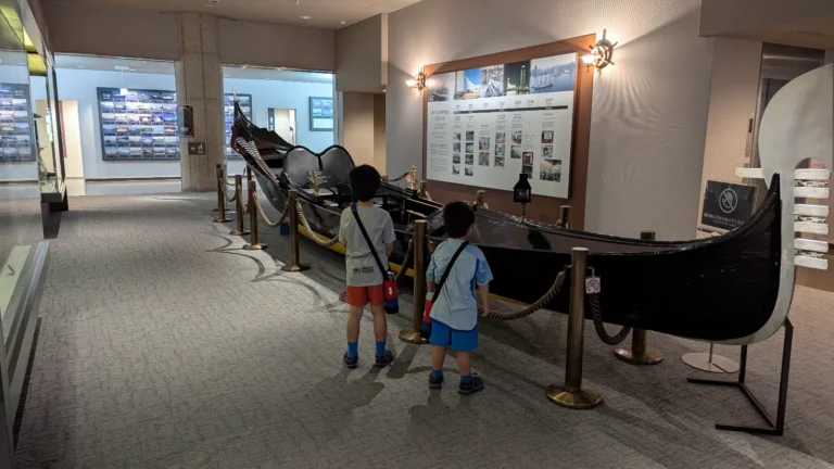 Luca & Nico observing a historic gondola exhibit inside the Kobe Maritime Museum, part of a family-friendly visit in Kobe, Japan.