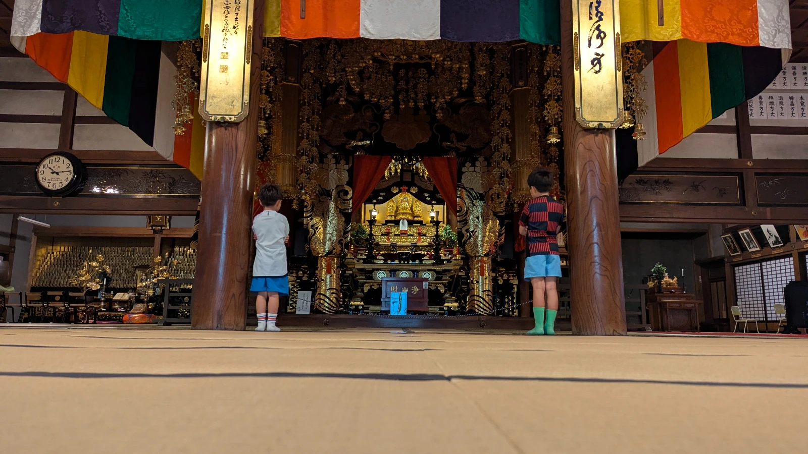 Two children quietly observe the ornate golden altar inside Kōryūji Temple in Hakodate, surrounded by colorful banners and traditional decor.