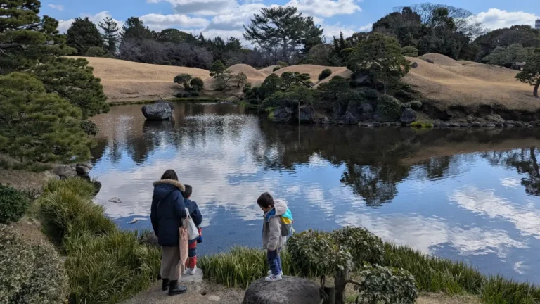 Luca & Nico, and Mama stand beside the tranquil pond at Suizenji Jojuen Garden in Kumamoto, Japan during a quiet moment on a family adventure.