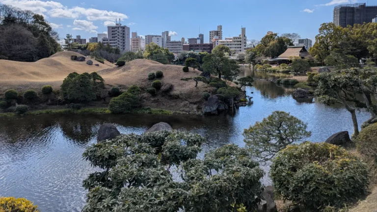 A scenic view of Suizenji Jojuen Garden in Kumamoto, with manicured hills, a traditional tea house, and a peaceful pond; one of the best places to visit in Kumamoto with kids for a relaxing nature walk and cultural experience.