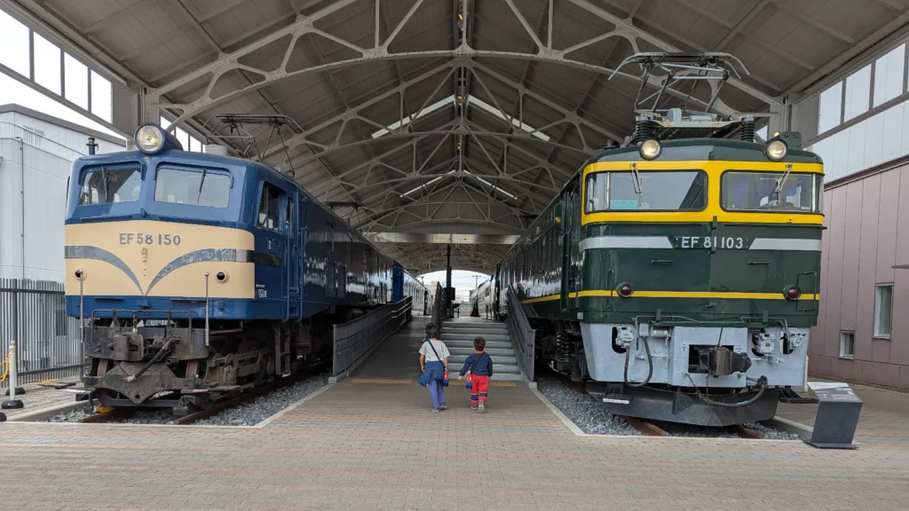 Luca & Nico walking between vintage trains at the Kyoto Railway Museum’s outdoor train yard, showcasing classic Japanese railroad engines.