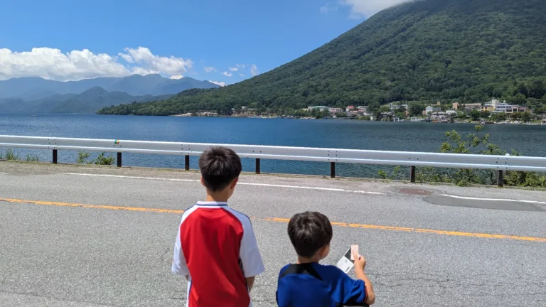 Two kids standing along the road overlooking Lake Chuzenji in Nikko, Japan, with Mount Nantai in the background — a family-friendly sightseeing spot.