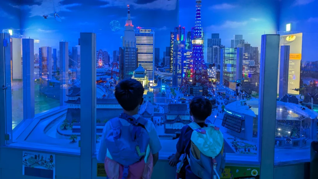 Luca & Nico with backpacks looking at Miniland Tokyo inside Legoland Discovery Center Tokyo in Odaiba, featuring Lego Tokyo Tower at night.