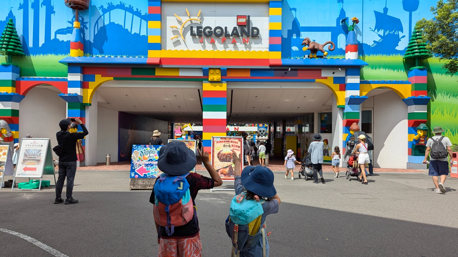 Luca and Nico, in sun hats, wave at the brick-striped main entrance of LEGOLAND Japan in Nagoya.