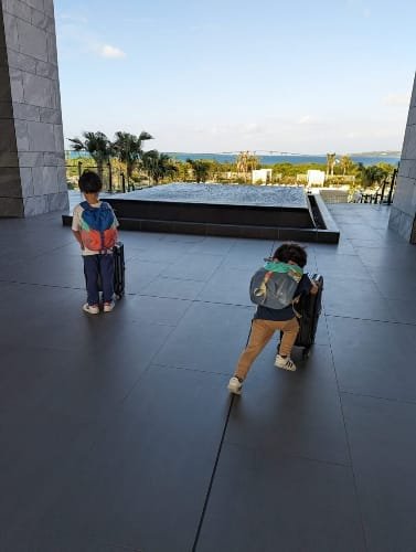 Two young children wearing backpacks walk on a sleek gray tiled floor, one pulling a small suitcase. They are in an open area with modern architecture, overlooking a scenic view of palm trees, a reflecting pool, and the ocean in the background under a clear blue sky at the Hilton Miyakojima, also known as Miyako Island.