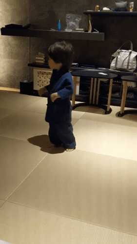 A young child in a navy yukata (traditional Japanese robe) stands barefoot on tatami flooring, exploring a serene and minimalist Japanese room.