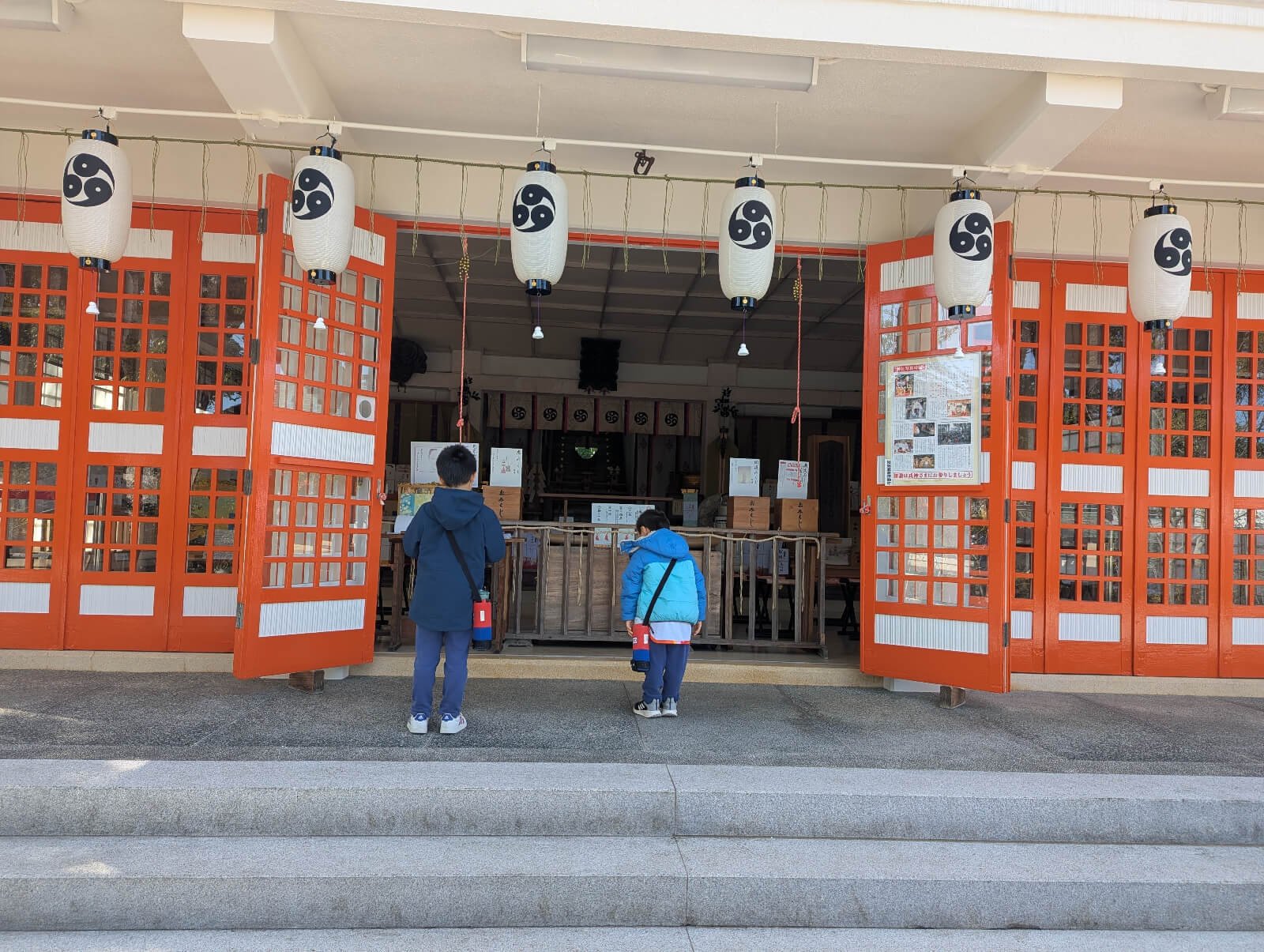 Two children dressed in warm jackets stand at the entrance of a traditional Japanese shrine, gazing inside with curiosity. The shrine features bright orange wooden doors and white lanterns with a black tomoe symbol, creating a serene and culturally rich scene of family travel in Japan.