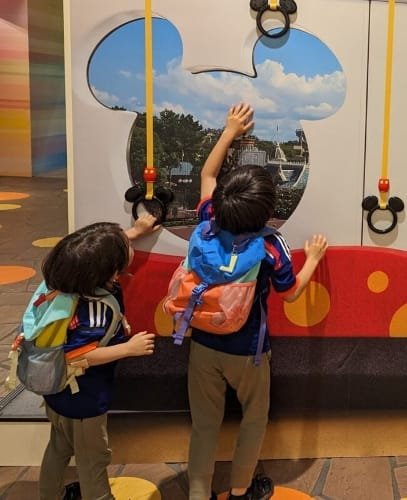 Luca & Nico dressed in colorful outfits with small Patagonia backpakcs, explore a playful display on a Tokyo Disney Resort train. They are reaching towards Mickey Mouse shaped windows with scenic images of the park and Cinderella's Castle in the background, accompanied by yellow handrails.