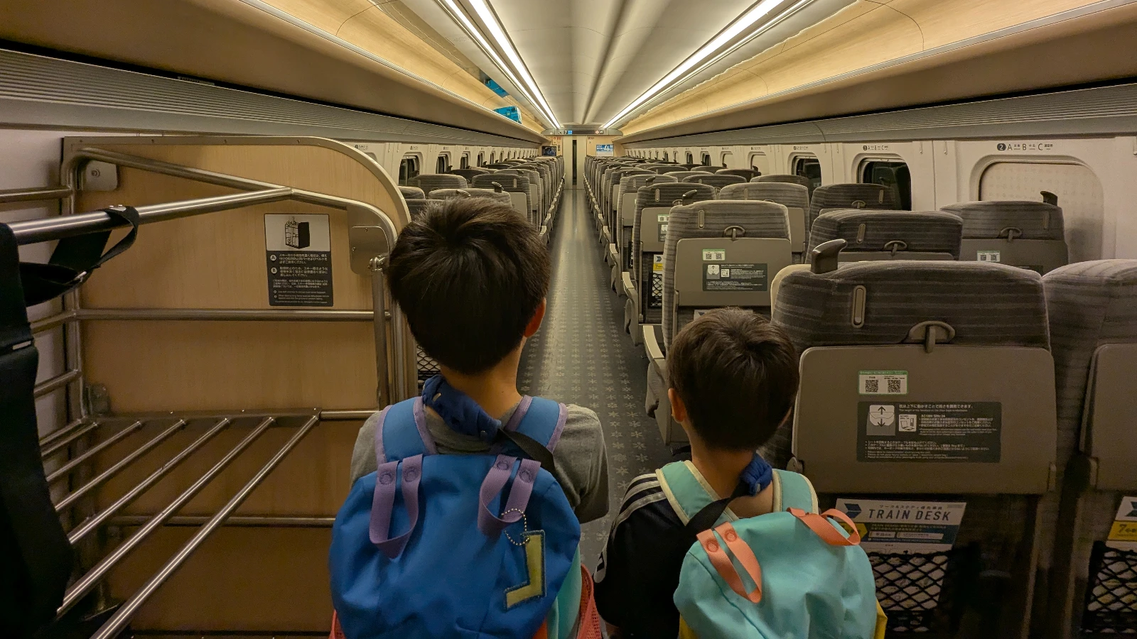 Luca and Nico standing inside a Shinkansen train car in Japan, wearing backpacks and facing a long aisle of empty seats with luggage racks on the side